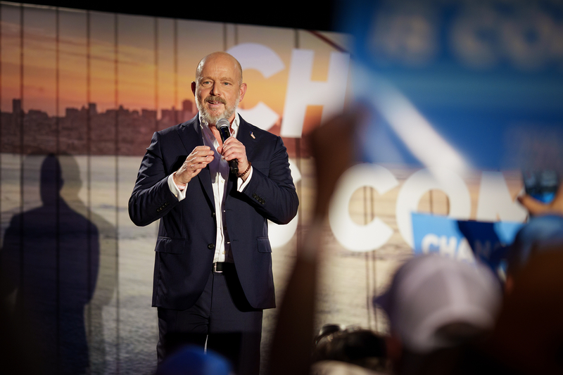 A person speaks into a handheld microphone on a stage, with supporters and campaign signs blurred in the foreground and a large backdrop behind them.