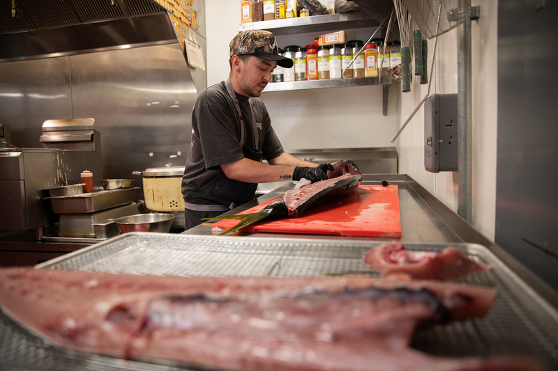 A person uses a small dish rag to clean off the remaining blood of a yellowtail cut open inside the kitchen of a restaurant. A separate belly piece of yellowtail can be seen in the blurry foreground.