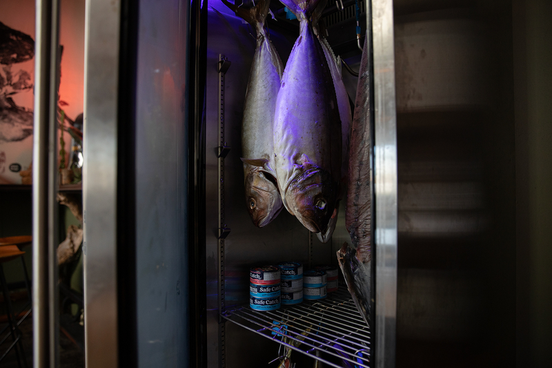 Aged yellowfish hand from the ceiling of a dry ager refrigerator illuminated by a soft purple light. Cans of tuna can be seen sitting on a shelf tiowards the bottom of the dry ager.