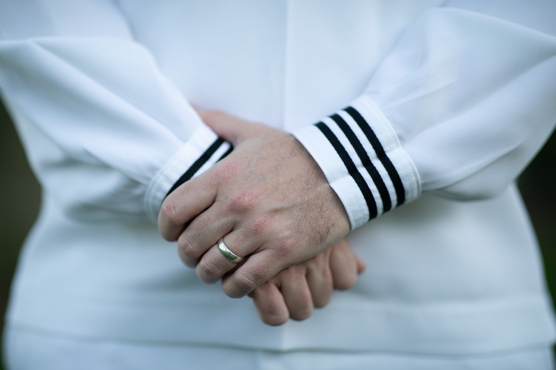 A close-up of a person's wedding ring on their left ring finger, as both hands rest on top of one another in front of their stomach while standing straight. The person wears a white Navy uniform with black details on it.