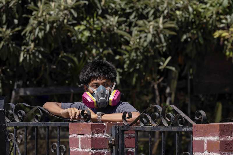 A person wearing a respirator leans on a wrought-iron gate while looking toward the left. Trees fill the background, and the person appears to be observing activity outside the frame.