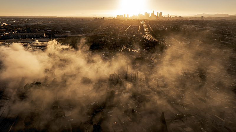 An aerial view shows thick smoke drifting over an industrial neighborhood as the downtown Los Angeles skyline glows in the distance at sunset. The haze blankets homes, warehouses and streets below, with sunlight filtering through the smoke in golden beams.