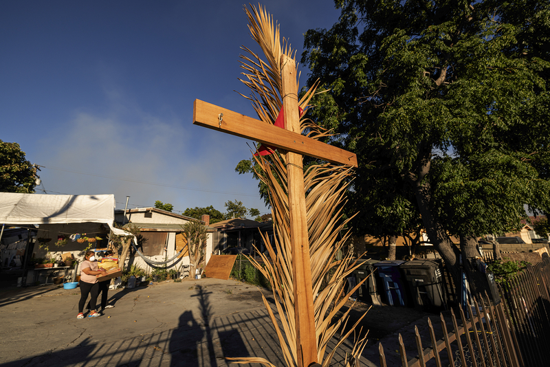 A wooden cross wrapped with dried palm fronds stands in the foreground outside a home as dark smoke fills the sky in the distance. In the yard behind it, two people carry boxes while a white canopy, trees and neighboring houses are bathed in warm evening light.
