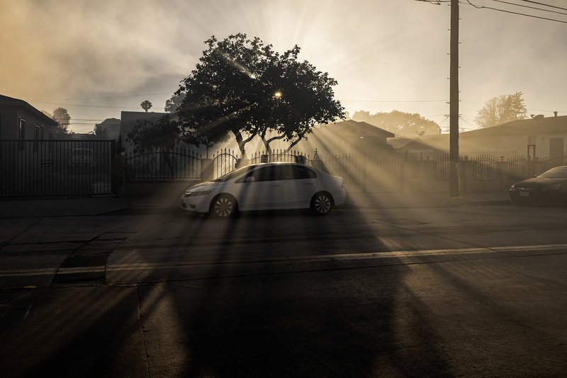 Sunlight streams through thick smoke over a residential street, creating dramatic rays around a large tree and parked white car. The hazy air softens nearby homes, utility poles and fences as smoke from a nearby warehouse fire drifts through the neighborhood.