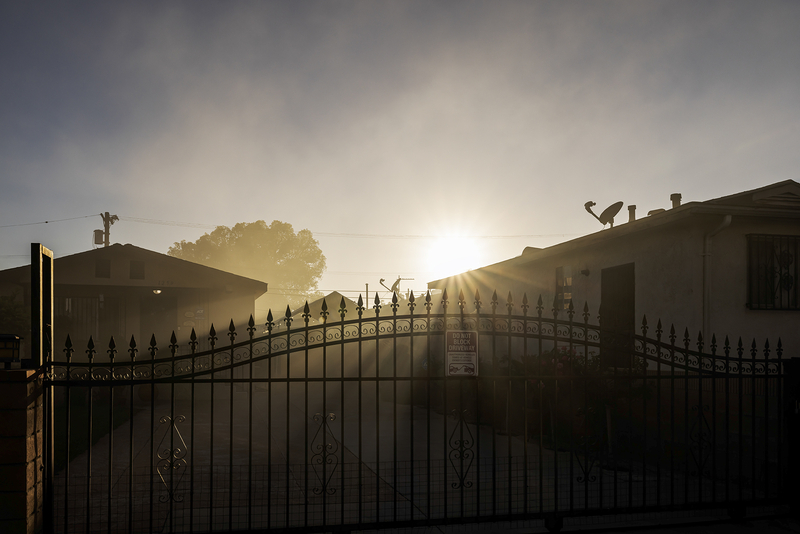 The sun rises through smoke hanging over a residential neighborhood, casting golden rays between two homes and across a wrought-iron driveway gate. The hazy air softens the outlines of nearby trees and rooftops, reflecting smoke from a nearby warehouse fire.