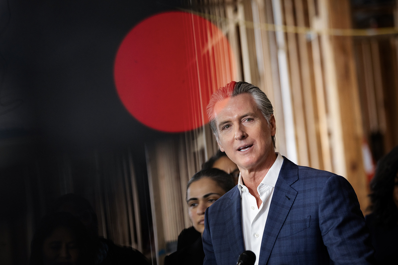 California Gov. Gavin Newsom wears a blue suit jacket and white shirt while speaking indoors, with several people standing behind him and a large red circular light in the foreground.
