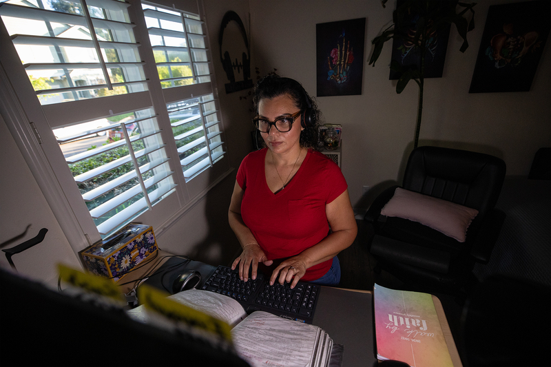A person wearing a red shirt and a headset sits in front of a desk and types into a keyboard, in a dimly lit room with a window overlooking a residential street.