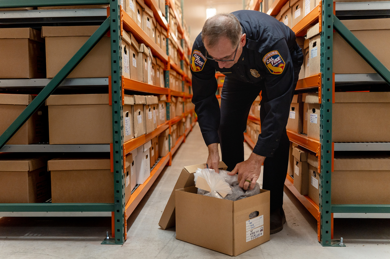 A man in a Cal Fire uniform bends over to open a cardboard box on the ground, revealing that it is full of seeds.
