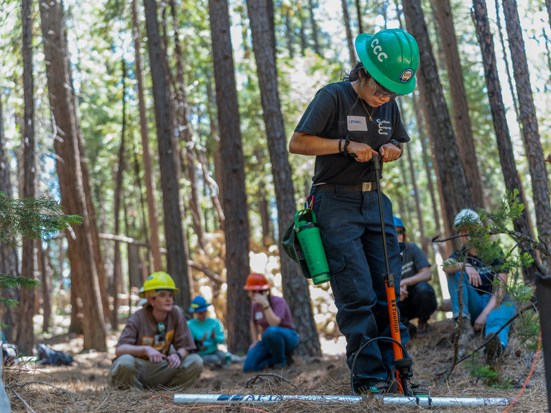 A woman in a hard hat pumps air into a tool used to launch rope as other people in hard hats watch on from a dense forest.