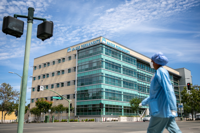 A person dressed head to toe in blue scrubs looks towards a Kaiser Permanente as they walk across an intersection.