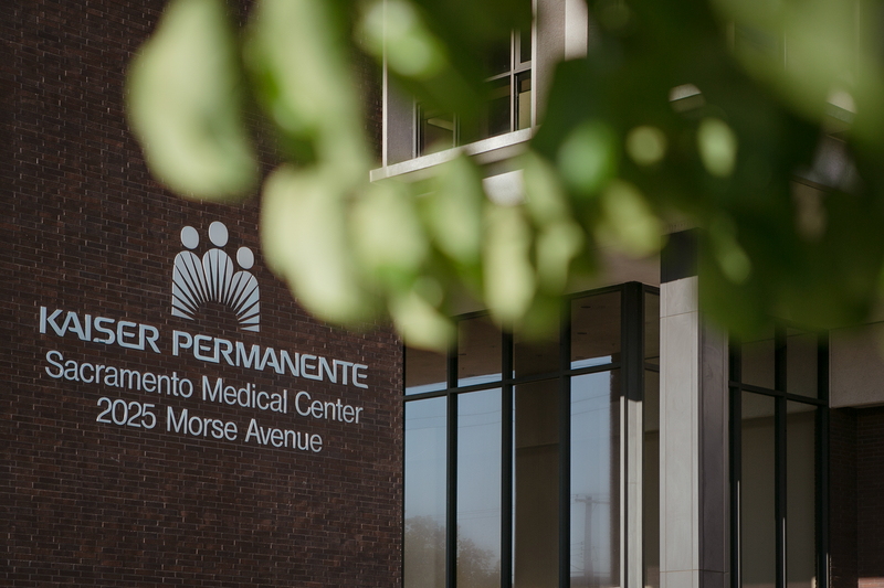 The Kaiser Permanente logo with the location address is displayed on a brick building. Leaves from a nearby tree can be seen in the blurred foreground.
