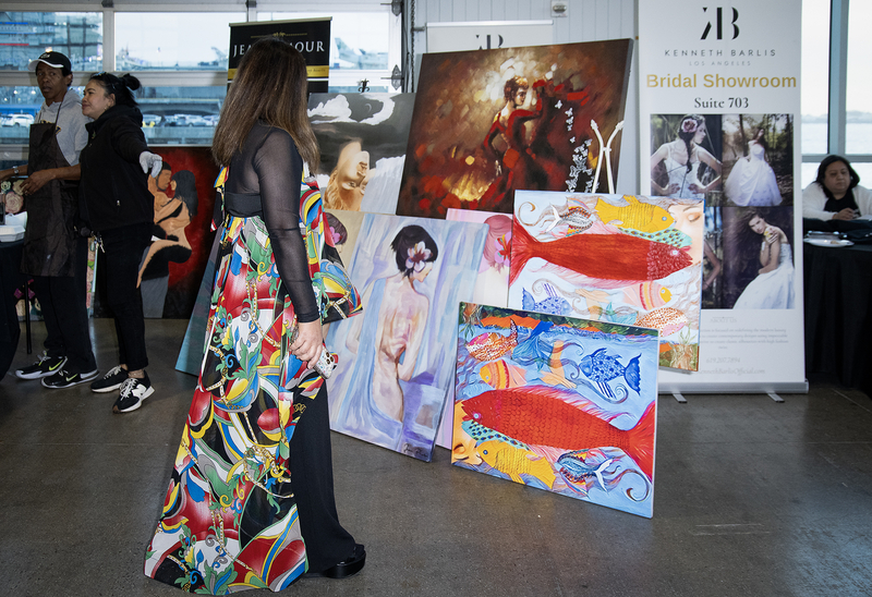 A person in a colorful patterned dress looks at a display of paintings, including portraits and brightly colored fish, at an indoor art exhibition with vendors and attendees in the background.