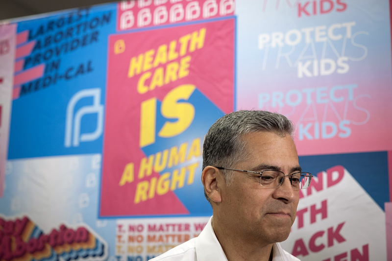 Xavier Becerra stands in front of a colorful wall display with messages about health care, reproductive rights and protecting trans kids.