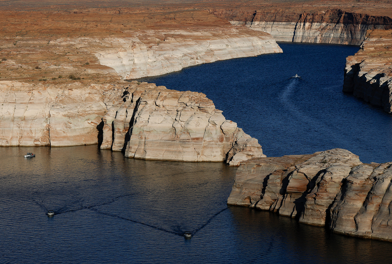 An elevated view looks over a narrow stretch of Lake Powell winding between layered sandstone cliffs marked by broad white mineral stains from higher water levels. Several small boats travel through the deep blue water, emphasizing the reservoir's low level against the expansive desert landscape.