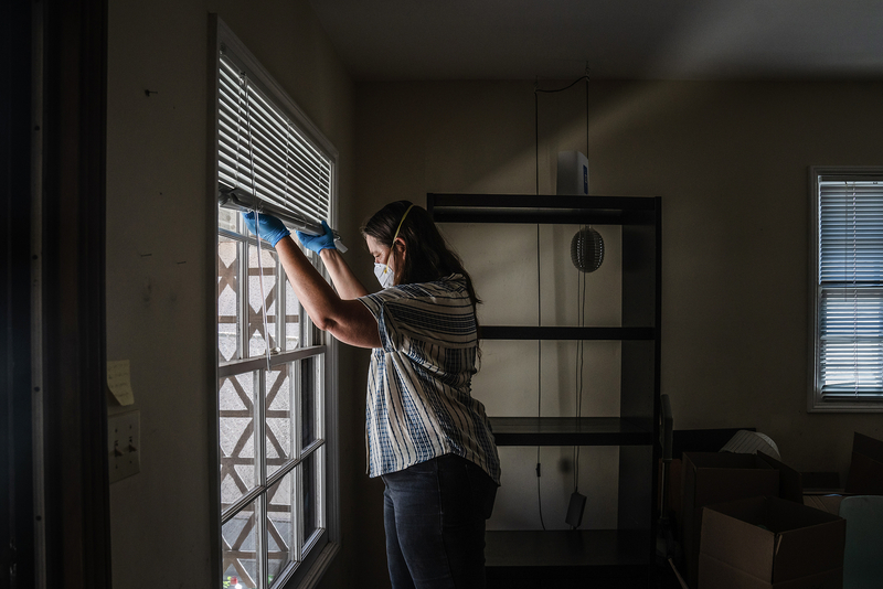 A person wearing a face mask and blue gloves stands inside a sparsely furnished apartment, lifting the blinds to look out a window. Empty shelves and moving boxes are visible in the dimly lit room behind them.