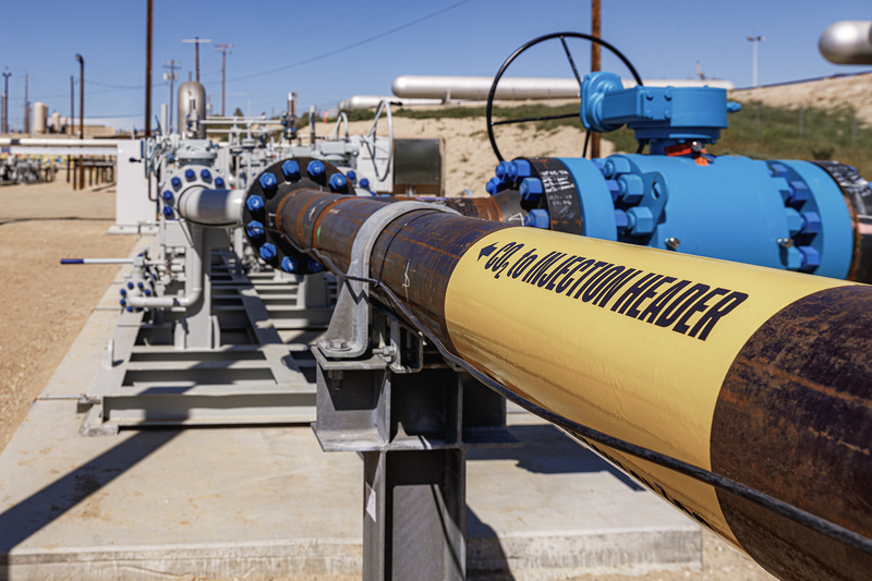 A close-up view looks down a pipeline fitted with blue valves and industrial equipment at an energy facility. A yellow sleeve wrapped around the pipe is labeled “CO₂ INJECTION HEADER,” indicating infrastructure used to transport carbon dioxide for underground injection.