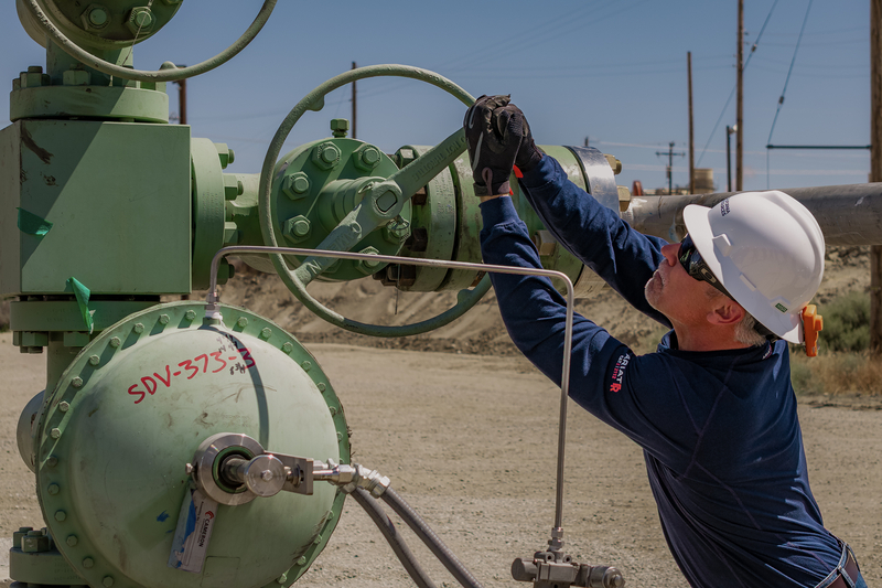 A worker wearing a white hard hat, safety glasses and gloves adjusts a large green valve at an industrial carbon dioxide injection facility. He reaches up to turn the handwheel on the pipeline equipment, with utility poles and industrial infrastructure visible in the background.