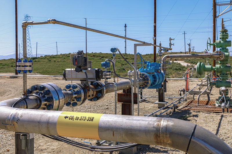 A network of pipes, valves and monitoring equipment at a carbon capture facility sits in a dry, open landscape. A yellow label on a large foreground pipeline reads "CO₂ to 373-35R."