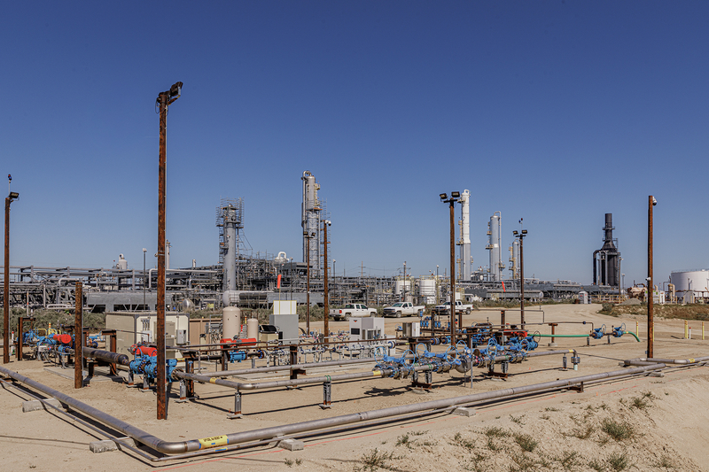 A large industrial carbon capture and storage facility with an extensive network of pipes, valves and wellhead equipment stretches across a dry landscape, with refinery towers and processing units rising in the background beneath a clear blue sky.