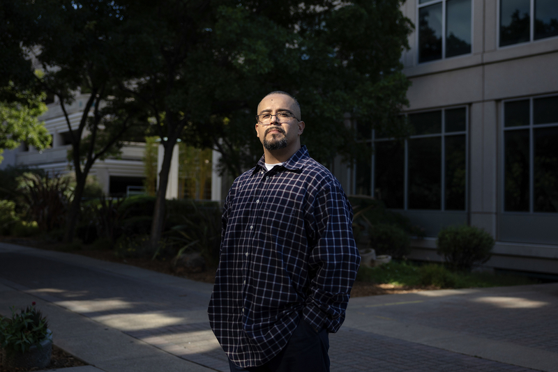 A person stands outside an office building, wearing glasses and a dark plaid button-down shirt with their hands in their pockets. Sunlight illuminates their face and upper body while trees and the building behind them remain largely in shadow.