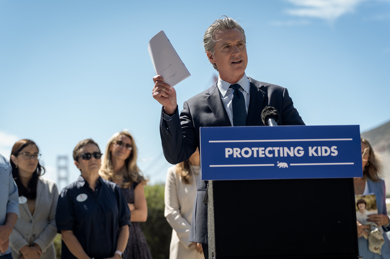 Gov. Gavin Newsom holds up documents while speaking at a podium with a “Protecting Kids” sign during a press conference at the Bay Area Discovery Museum in Sausalito. Several people stand behind him.