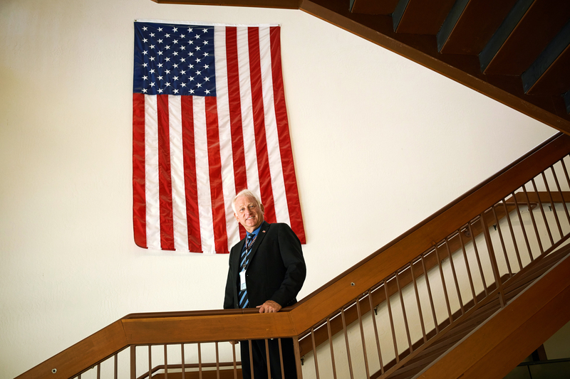 A man in a suit and tie stands on a staircase in front of an American flag hanging on the wall.