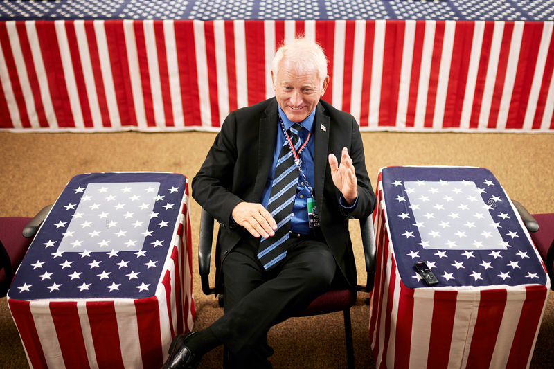 A man in a suit and tie uses hand gestures as he speaks in a room where all surfaces are covered with American flag tablecloths.