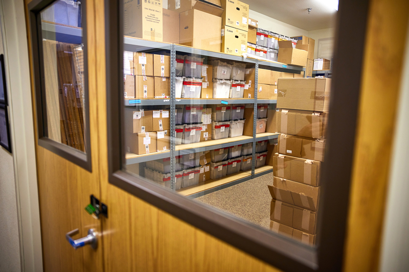 Shelves of cardboard and plastic boxes can be seen through a window on a set of locked and sealed doors.