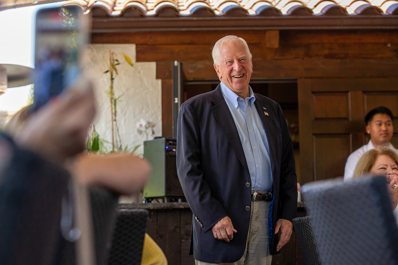 An elderly person wearing a blue blazer and gray slacks, laughs while standing in front of a restaurant filled with attendees. A person taking video with their phone can be seen in the blurry foreground.