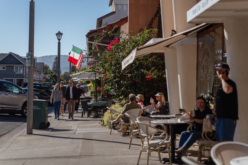People sit the outside sitting area of a restaurant overlooking a street with parked cars, as a small group of people walk by in the distance.