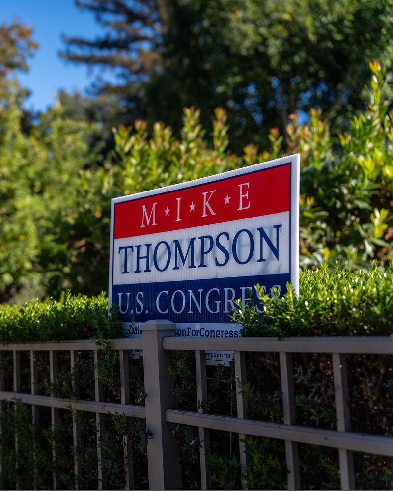 A red, white and blue campaign sign that reads, " Mike Thompson for U.S. Congress" is posted on a small bush next to fence in front of a home.
