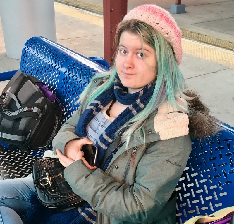A young adult sitting on a blue bench as they look up at a person taking a photo of them. The young adult has light blue and blonde hair and wears a pink crochet beret.