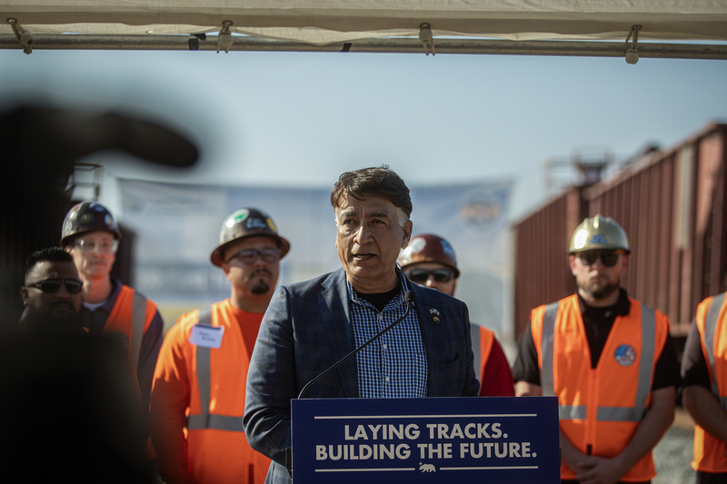 A man in a dark blazer speaks at a podium reading “Laying Tracks. Building the Future,” surrounded by workers wearing orange safety vests and hard hats at a rail yard.