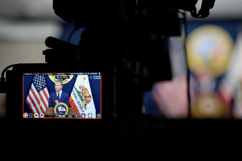 Gov. Gavin Newsom speaks at a podium between American and California state flags, seen through the monitor of a television camera during a press conference. The camera equipment is silhouetted in the foreground.