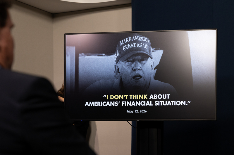 A television screen displays a black-and-white image of President Donald Trump wearing a “Make America Great Again” hat, alongside a quote reading, “I don’t think about Americans’ financial situation.” A person stands out of focus in the foreground.