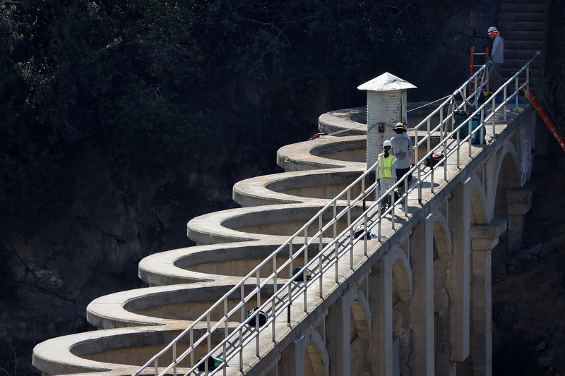 Three workers wearing hard hats stand along a narrow walkway with metal railings atop a concrete dam. A series of curved spillway structures extends alongside the walkway, above large concrete arches.