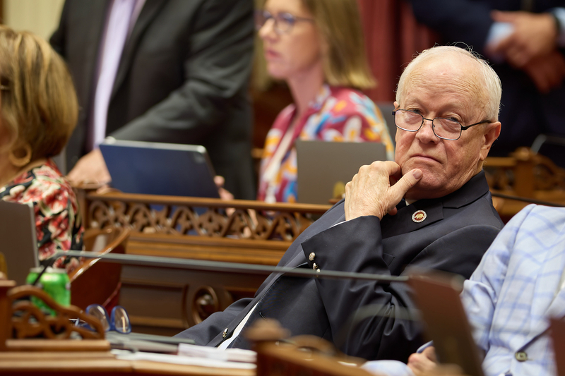 A lawmaker sits in front of a desk with their left hand resting on their chin as they look off into the distance. They are surrounded by other lawmakers sitting at their desks.