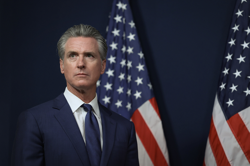 A person wearing a blue suit stands in front of two American flags while as they look straight off into the distance during a press conference.