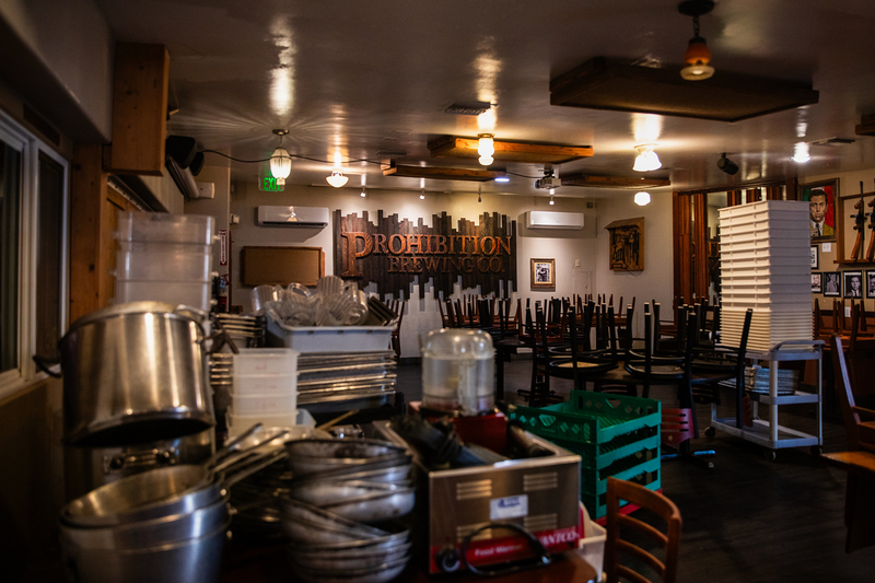 The dining area of a brewery with stacked dishes and kitchen equipments and chairs stacked on top of tables.