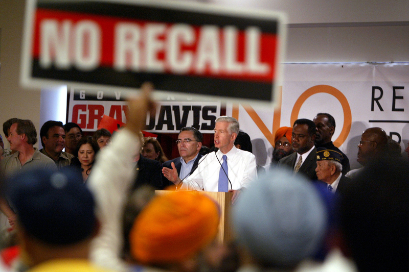 A person speaks to a crowd while standing behind a podium and surrounded by other people on stage. The crowd, come who hold signs that read, "No Recall" can be seen in the blurry foreground.