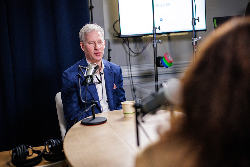 A person with short white hair speaks into a microphone sitting on a table during an podcast interview.
