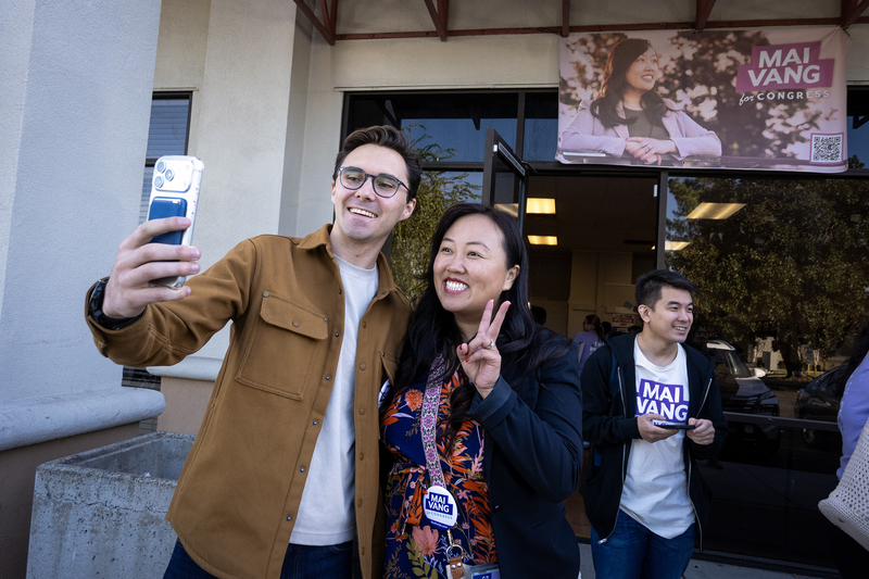 Congressional candidate Mai Vang smiles and flashes a peace sign while posing for a selfie with a supporter outside a campaign event, beneath a large Mai Vang for Congress banner.