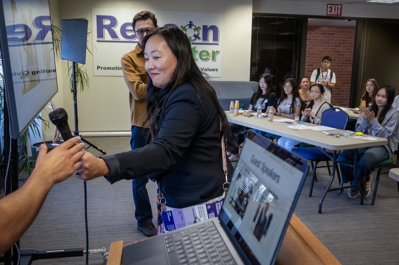 Congressional candidate Mai Vang accepts a microphone at an indoor campaign event as attendees seated at tables applaud.