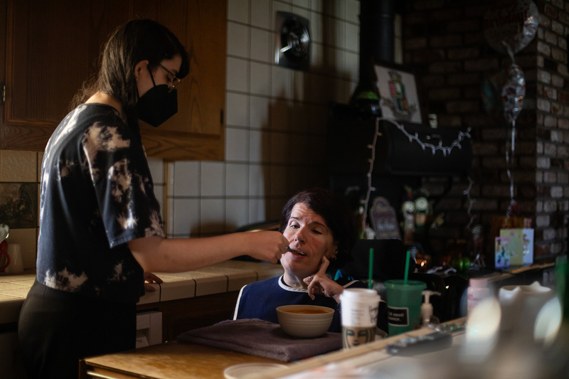 A person wearing a face mask, helps feed another person in a wheelchair.