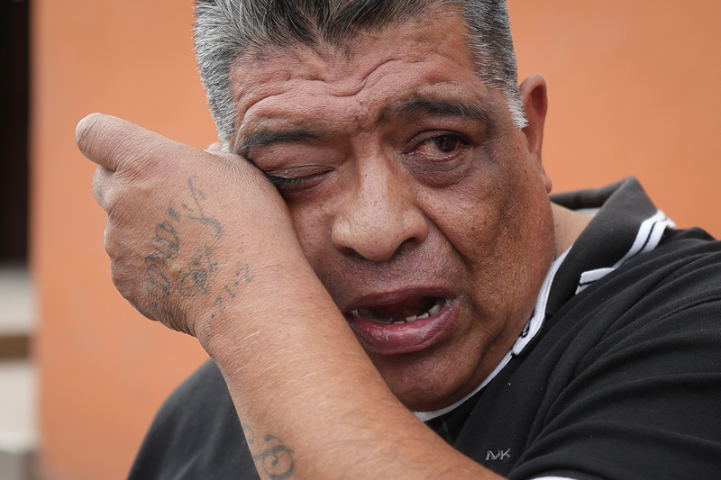 A close-up of Luis Arreguin wiping his eye with a tattooed hand. His mouth is open, and his face appears tearful against a blurred orange background.