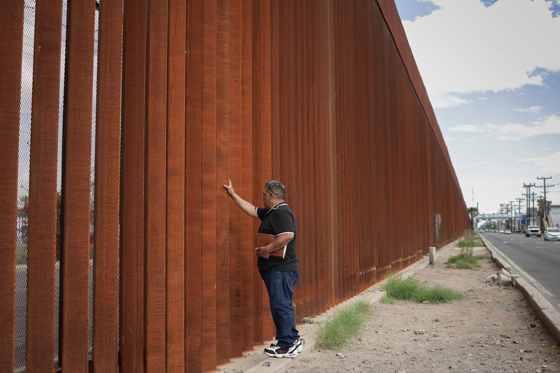 Luis Arreguin touches a tall, rust-colored steel border wall while holding a Bible under his arm. The wall stretches into the distance alongside a road.