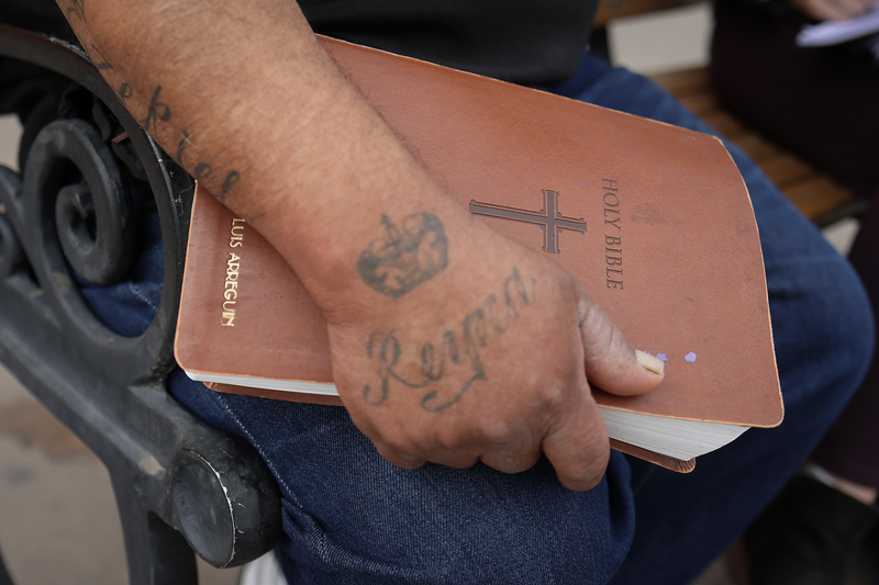 A close-up of Luis Arreguin’s tattooed hand resting on a brown leather-bound Bible in his lap. The Bible’s cover features an embossed cross and his name in gold lettering.