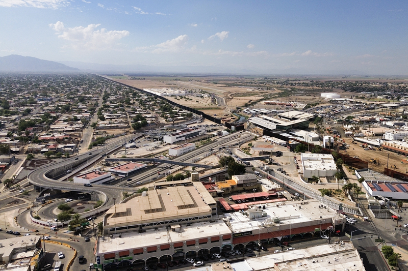 An aerial view of the U.S.-Mexico border crossing between Calexico and Mexicali, with roads, inspection facilities and neighborhoods surrounding the border wall.