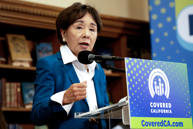 U.S. Rep. Doris Matsui speaks into a microphone at a podium displaying a Covered California sign, gesturing with one hand in front of wooden bookshelves.