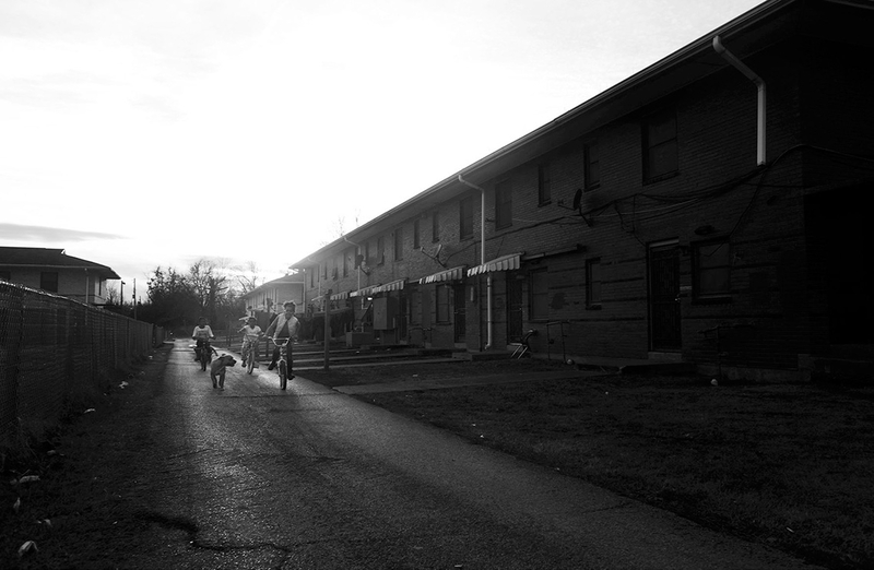 Three children ride bicycles next to a large apartment building. A dog runs next to them.